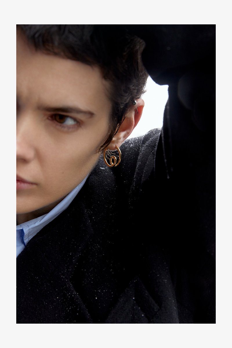 Boucle d'oreille en or avec un design tordu. Le modèle la porte avec un manteau noir. L'arrière-plan montre un cadre lumineux, légèrement texturé.
