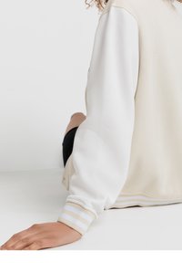Lekka bluza w kolorze kremowym i białym, z paskowanymi ściągaczami. Gładka tekstura, luźny krój i zamek błyskawiczny.