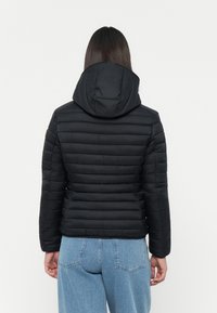 Zwarte pufferjack met horizontaal gestikte ontwerp, capuchon en ribgebreide manchetten. Gecombineerd met hooggehaalde blauwe jeans.