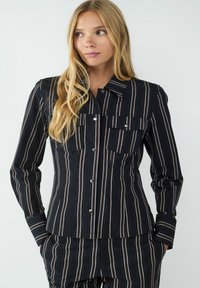 Schwarz gestreiftes Hemd mit zwei Brusttaschen, Knopfleiste und langen Ärmeln, das aus einem strukturierten Stoff mit horizontalen und vertikalen Streifen besteht.