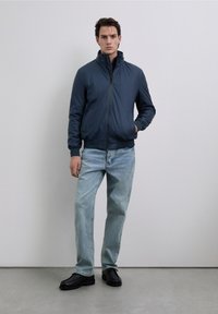 Marinfärgad bomberjacka med dragkedja fram, ribbade muddar och fåll, bärs tillsammans med ljusblå denimjeans och svarta skor. Minimalistisk design, slät textur.