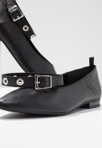 Chaussures plates noires à bout pointu avec brides réglables à la cheville comportant des boucles argentées et des œillets sur une surface blanche.