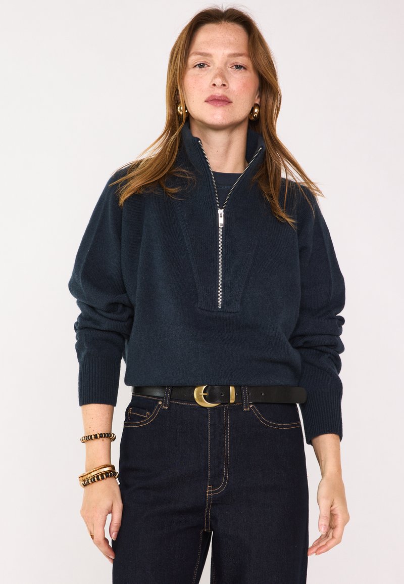 Maglione zip-up blu navy con colletto alto, polsini a coste e un motivo testurizzato. Indossato sopra una maglia nera e abbinato a jeans scuri e una cintura.