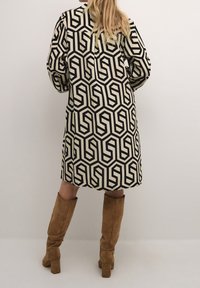 Mekko pitkillä hihoilla, jossa on geometrinen musta ja kerma patterni, löysä istuvuus ja pyöreä kaula-aukko. Yhdistetty vaaleanruskeisiin mokkanahkaisiin polvipitkiin saappaisiin.