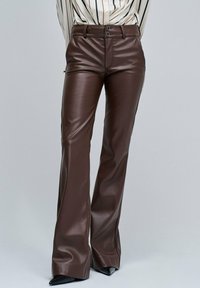 Hoge taille bruine leren broek met dubbele knoopsluiting, gedragen met zwarte puntige schoenen en een ingestopt gestreept shirt.