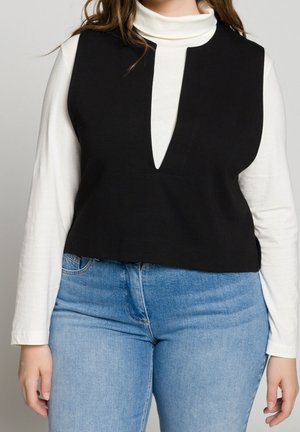 Femme portant un haut noir sans manches avec un décolleté en V profond sur un col roulé blanc et un jean bleu clair, debout devant un fond uni.
