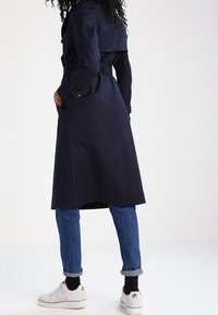 Kvinna som bär en marinblå trenchcoat, blå uppvikta jeans, svarta strumpor och vita Nike Air-sneakers, står med händerna i fickorna på trenchcoaten.