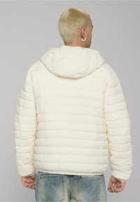 Pufferjakke i myk kremfarge med quiltet design og hette. Har lange ermer og ribbestrikkede mansjetter, kombinert med lyseblå denimbukser.