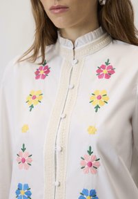 Bela bluza z barvnim cvetličnim vezenjem v roza, rumeni in modri barvi. Okrasni čipkast rob in podrobnosti gumbov obogatijo oblikovanje.