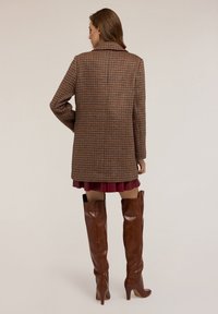 Abrigo de cuadro marrón con un corte relajado, que presenta un cuello con muescas y mangas largas. Combinado con una falda burdeos y botas marrones hasta la rodilla.