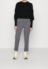 Person som bär en svart långärmad tröja, grå uppvikta byxor, vita ankelsockar och gula och vita sneakers mot vit bakgrund.