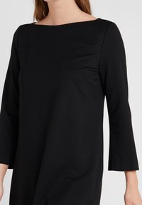 Robe noire à manches longues avec décolleté carré, en tissu doux. Présente un design simple sans motifs ni décorations visibles.