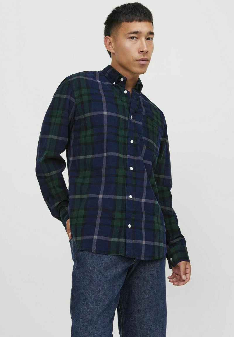 Camisa de cuadros en azul marino y verde, confeccionada en una tela suave. Presenta un cuello, mangas largas y botones blancos. Combinada con vaqueros azules.