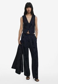 Navy floral-patterned vest top with a deep V-neck, paired with wide-leg trousers and a matching blazer, all in smooth fabric. Strappy sandals.