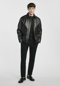 Czarna skórzana kurtka z pikowanymi detalami, z dwoma przednimi kieszeniami, noszona na czarnej koszuli, w zestawieniu z czarnymi spodniami i mokasynami.