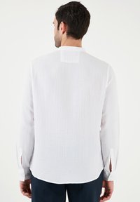 Camisa blanca de manga larga con tejido texturizado; cuenta con un escote redondo, puños con botón y un pequeño detalle de yugo en la parte posterior.