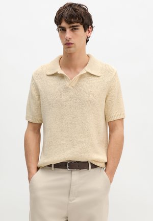 Jaunas vyras su tamsiais plaukais, dėvintis smėlio spalvos trumpomis rankovėmis megztą polo marškinėlį, smėlio spalvos kelnes ir tamsiai rudos spalvos diržą, rankos kišenėse.