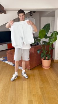 Muškarac u srebrnim cipelama i sivim šortsevima drži par bijelih šortseva u dnevnoj sobi s velikom posudom s biljkom i smešnim kaučem.