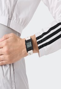 Asmuo, vilkintis baltą striukę su juodomis juostelėmis, demonstruoja skaitmeninį laikrodį su baltu dirželiu ir minimalistiniu juodu ekranu.