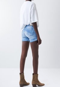 Světle modré džínsové šortky s třepeným lemem, v kombinaci s volně střiženým bílým tričkem a hnědými kotníkovými botami, viděné zezadu.