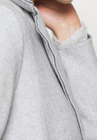 Gros plan d'une personne portant un manteau en laine gris clair avec une couture visible et une main touchant son cou.