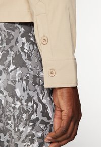 Beige jakk, mille varrukas on nööbiga kinnitamine, tekstureeritud kangas, koos musterhallide pükstega, millel on abstraktne disain.