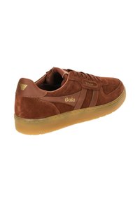 Wildlederbraune Sneaker mit Gummisohle, ausgestattet mit flachen Schnürsenkeln, goldener Markenprägung und seitlichen Streifen. Abgerundete Zehenpartie und gepolsterter Kragen für zusätzlichen Komfort.