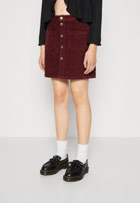 Falda de pana burdeos con cintura ajustada, cierre de botón en el frente y dos bolsillos grandes, combinada con zapatos negros y calcetines blancos.