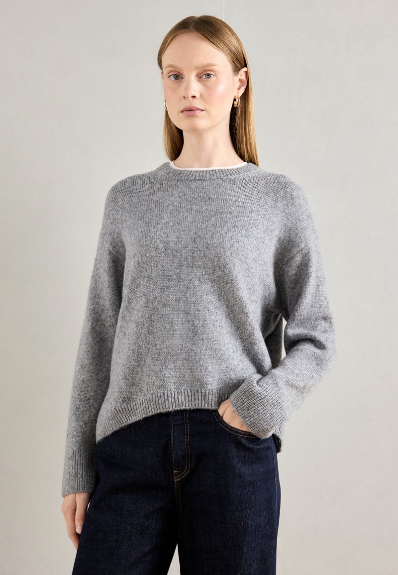 Mulher com um suéter de malha cinza e jeans azul escuro, de pé com uma mão no bolso, contra um fundo claro e liso.