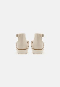 Beige Riemchensandalen mit einem glatten Lederobermaterial, offenem Design, flacher Sohle und verstellbaren Schnallen am Rücken.