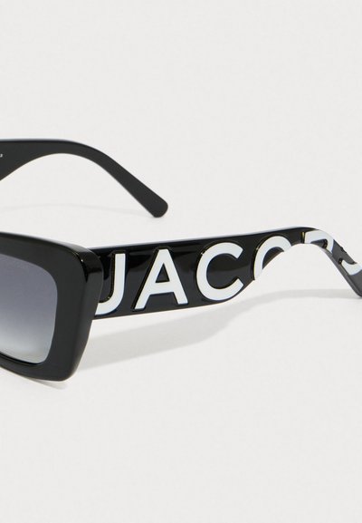 Czarne prostokątne okulary przeciwsłoneczne z gradientowymi soczewkami i odważnym białym napisem "JACOBS" na ramieniu przeciwko prostemu tłu.