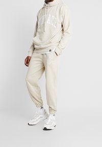 Beige huvtröja med vitt logotyp, ficka fram; matchande beige sweatpants med logodetalj; ihop med vita sneakers och strumpor.