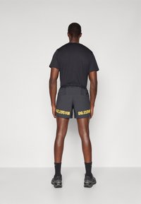 Czarny sportowy t-shirt zestawiony z ciemnymi szortami z żółtym napisem "42,195 km" i "26,219 mi", noszony z czarnymi skarpetkami i butami.
