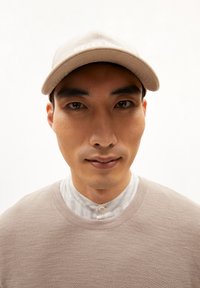 Cappellino da baseball beige con ricamo bianco, maglione a trama beige chiaro e camicia a righe con colletto, il tutto su uno sfondo neutro.