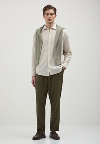 Hellbeige Hemd mit Knöpfen, olivgrüne Hose, grüner Strickpullover über die Schultern drapiert, braune Schuhe, glatte Stofftexturen.
