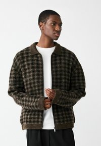 Brūns rakstains rāvējslēdzēja jaka ar tumšu apkakli, ribotām piedurknēm un mīkstu tekstūru. Uzvilkts virs baltas krekla ar brīvu piegriezumu.