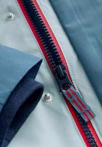 Lyseblåt jakke stof med en navy- og rød lynlås. Har sølvfarvet trykknap og blå ribstrikket manchet. Teksturerede detaljer og rene linjer.