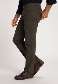Pantalones verdes olivo de corte slim, con bolsillos laterales y traseros, combinados con zapatos de cuero marrón con cordones, destacando la textura de la tela.