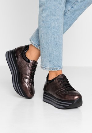 Osoba nosząca metaliczne ciemnobrązowe sneakersy na platformie z czarnymi sznurówkami i paskowanymi grubymi podeszwami, w połączeniu z jasnoniebieskimi dżinsami.