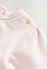 Vaaleanpunainen puuvillapaita, jossa on metallinhohtoinen kultainen reuna kaula-aukolla ja pieni kultainen sydämenmuotoinen yksityiskohta.