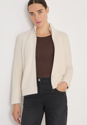 Frau trägt eine hellbeige, flauschige Zip-Jacke, ein dunkelbraunes Oberteil und schwarze hochgeschnittene Hosen, mit einer Hand in der Tasche.