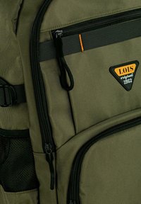 Mochila color verde oliva hecha de un tejido texturizado, con cremalleras negras, bolsillo de malla, costuras naranjas y un parche de logo en la parte delantera.