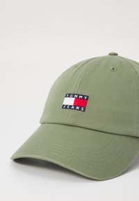 Olíva zöld baseball sapka ívelt peremmel; téglalap alakú, hímzett logóval rendelkezik tengerészkék, fehér és piros színekben, melyen az "TOMMY JEANS" felirat olvasható.