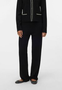 Cardigan noir en maille avec bordure blanche contrastante, boutonnage sur le devant et deux poches, associé à un pantalon noir ample assorti et des ballerines noires à motifs.