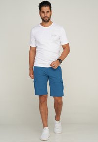 Weißes kurzärmeliges T-Shirt mit kleinem Logo, kombiniert mit blauen Cargoshorts mit zwei Fronttaschen und weißen Sportschuhen.