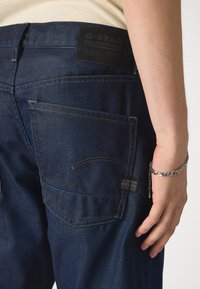 Vaqueros de mezclilla azul oscuro con un bolsillo trasero alto, parche de cuero de G-Star Originals y detalles de costura sutiles. Mano descansando en la cadera.