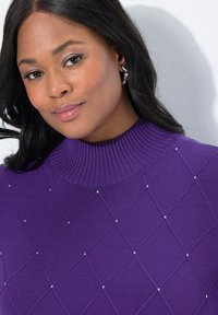 Žena s tmavými vlasy, která nosí stříbrné náušnice a fialový žebrovaný svetr s diamantovým vzorem a malými cvočky, na bílém pozadí.