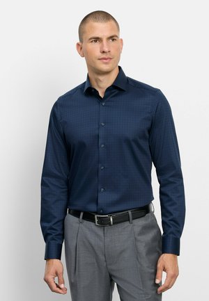 Uomo che indossa una camicia elegante aderente blu navy a maniche lunghe infilata in pantaloni eleganti grigi con cintura nera, in piedi e guarda di lato.