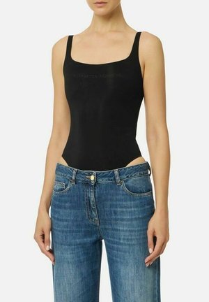 Donna che indossa un body nero senza maniche con scollo quadrato infilato in jeans blu con chiusura a bottoni su sfondo bianco.