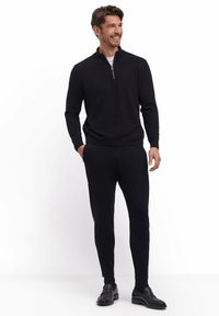 Schwarzer Zip-Hals-Pullover kombiniert mit schwarzen, eng anliegenden Hosen und schwarzen Schuhen. Der Stoff wirkt weich, hat eine glatte Textur und ein minimalistisches Design.